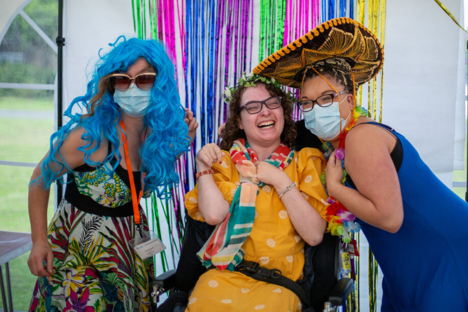 A young lady in a wheelchair laughing with two support workers wearing fancy dress. One lady wears a blue wig and the other a cowboy hat.