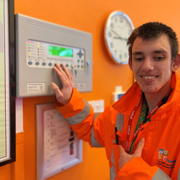 Student wearing orange hi-viz jacket smiling whilst operating the fire alarm panel