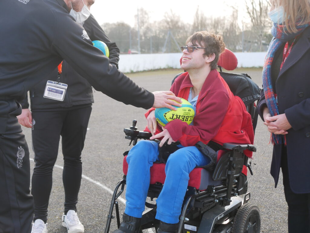 Young man in a wheelchair holding a rugby ball and smiling up at a coach