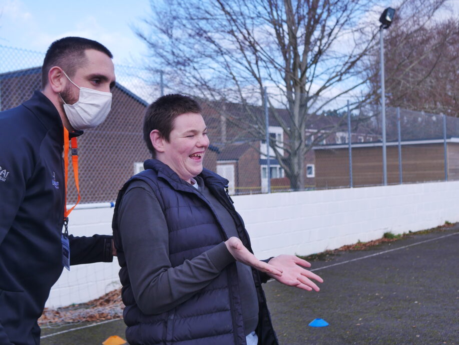 A young man is stood next to one of the community coaches. He is smiling and holding his hands out ready to catch a ball