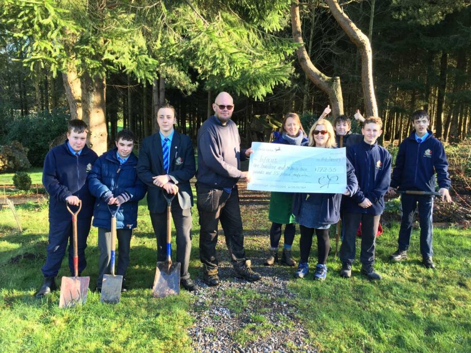 Pupils in school uniform holding gardening tools and standing in a line with 2 adults holding a giant cheque. All are outside in a forest.