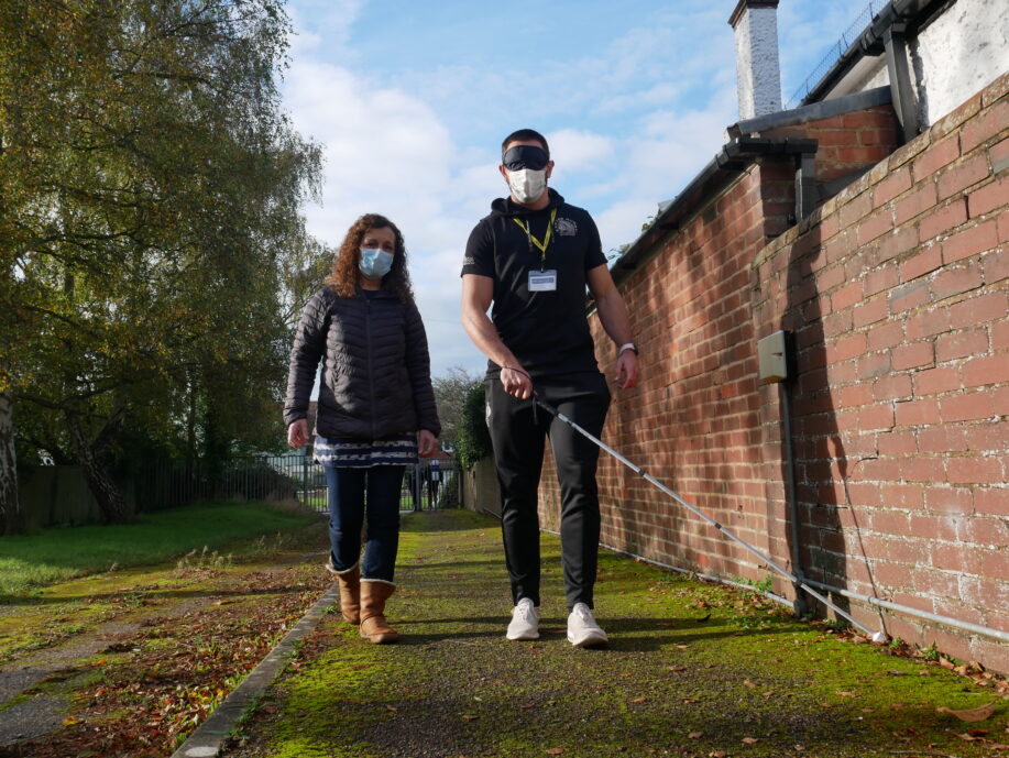 Man wearing a sleep shade and using a long cane. He's walking alongside a lady who is guiding him