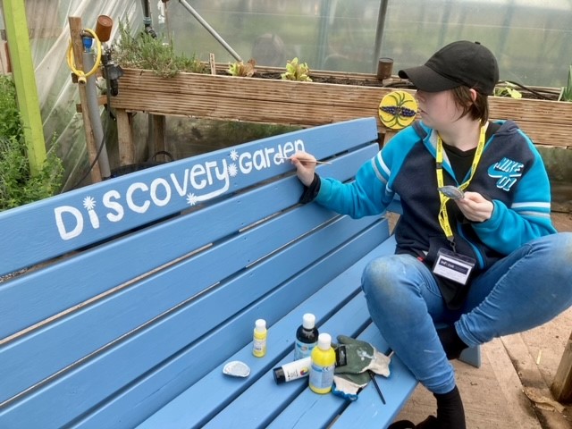 Painting the words Discovery Garden onto a blue garden bench