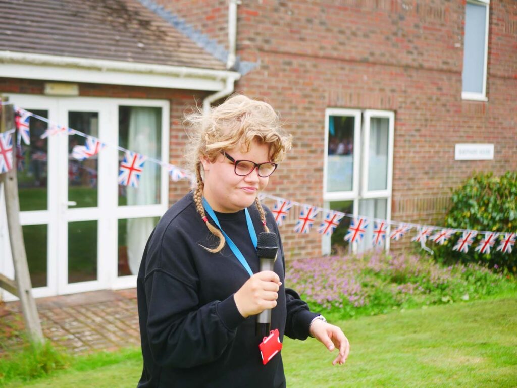 Young lady in glasses outside holding a microphone in front of a building and jubilee bunting