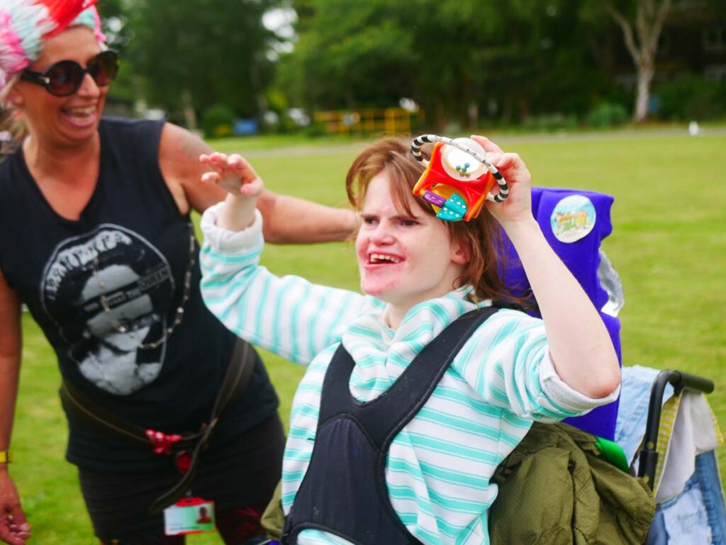 Young lady in a wheelchair smiling and holding up a toy with lady in a wig standing next to her.