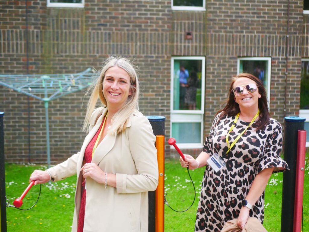 Two ladies outside in front of a building holding mallets and striking metal tubes.