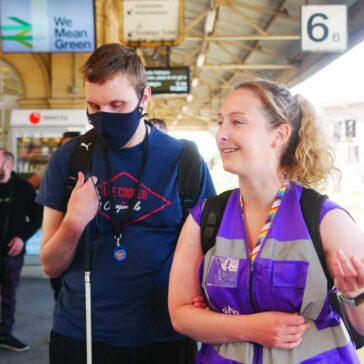 Young man in a face covering holding a long cane and being supported along the station platform by a lady in a purple vest.