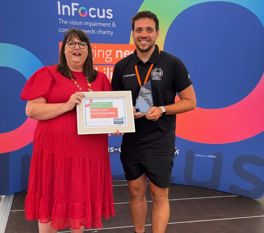 Jane Bell is standing in front of an infocus backdrop passing a certificate to Tom who is wearing black rugby kit