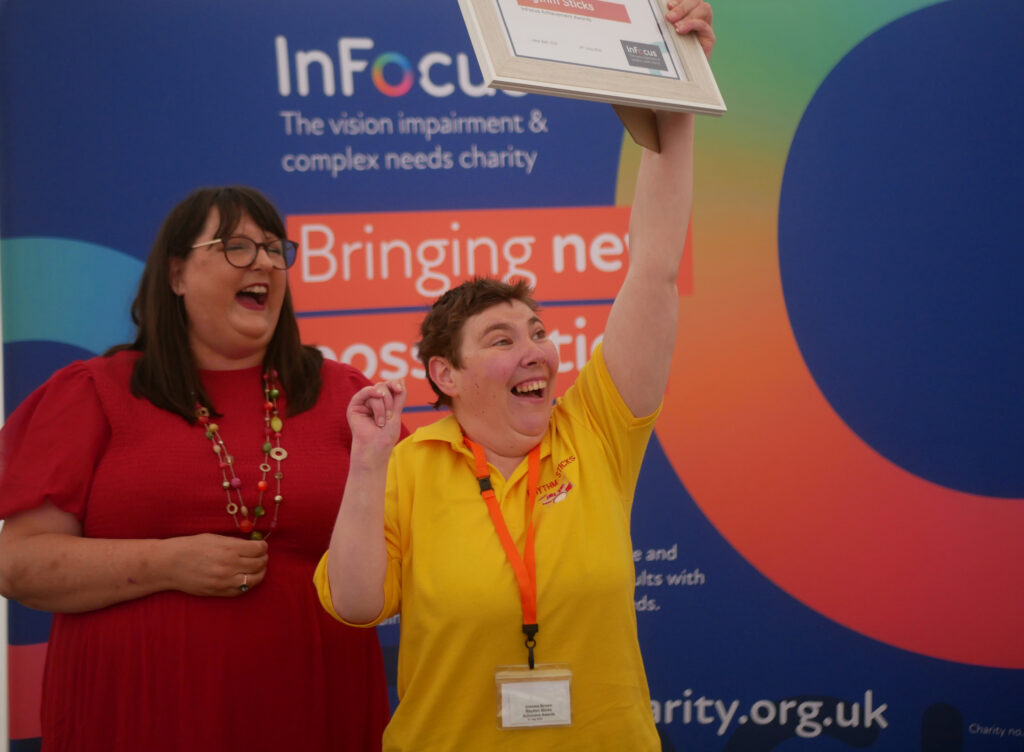 YOung lady in a yellow t shirt smiling as she holds a certificate high in the air. There's a lady in red laughing behind her and a bright blue banner