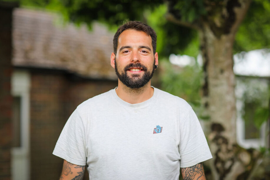 Picture of Ant standing outside a classroom at InFocus. He's wearing a white tshirt and looking at the camera