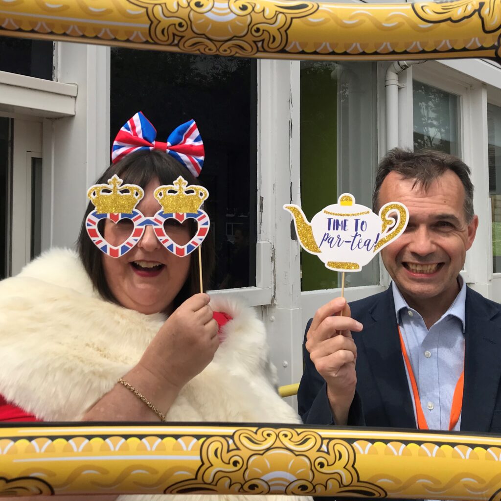 Two people posing for a photo through a blow up frame. One is holding up a cardboard tea pot and the other a pair of union jack cardboard glasses.