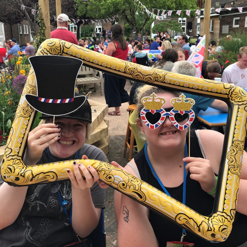 Two students posing for a photo through a blow up frame. One is holding up a cardboard bowler hat and the other a pair of union jack cardboard glasses.