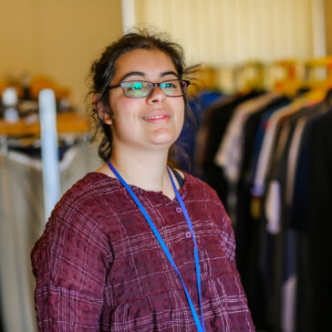 Young person looking away from the camera into the light. In the background there are clothes hanging from rails.