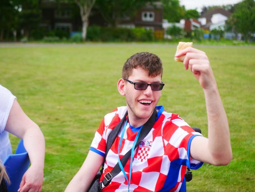 Young man in a wheelchair and red white chequed football shirt holding up a sausage roll
