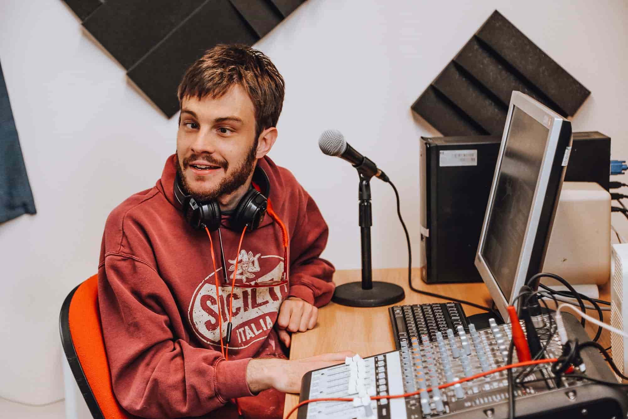 Young man sat at a computer with a microphone and sound mixing desk in front of him