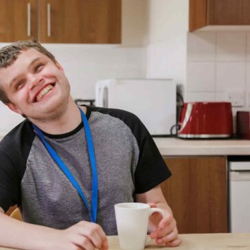 Young man with blue lanyard sitting in the kitchen with a cup of tea with a huge smile on his face