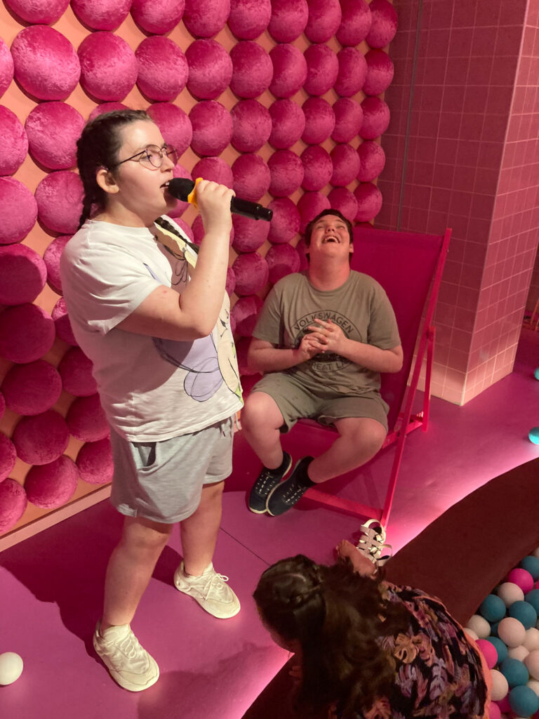 A young woman sings karaoke while a young man laughs next to her