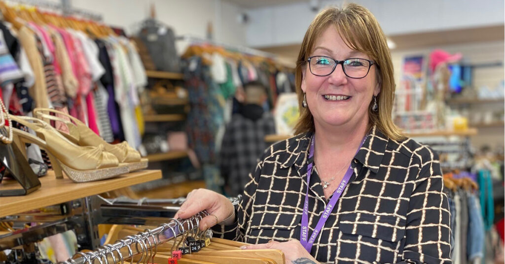 a lady smiling for the photo. She's standing in a charity shop