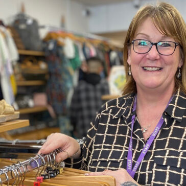 a lady smiling for the photo. She's standing in a charity shop