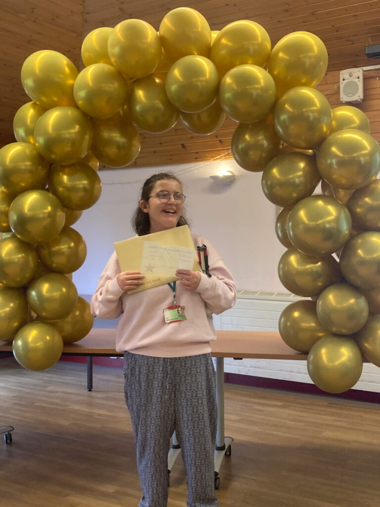 A smiling student holding a large envelope under an arch of golden balloons. 