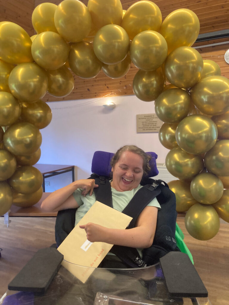 A smiling student holding a large envelope under an arch of golden balloons. 