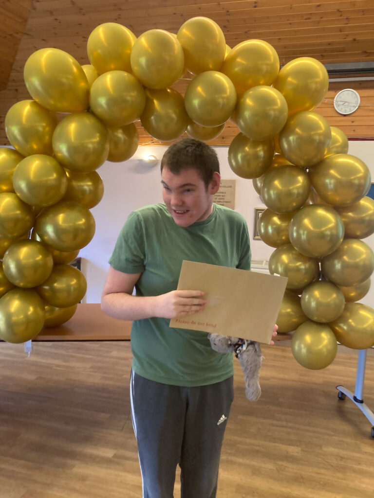 A smiling student holding a large envelope under an arch of golden balloons. 