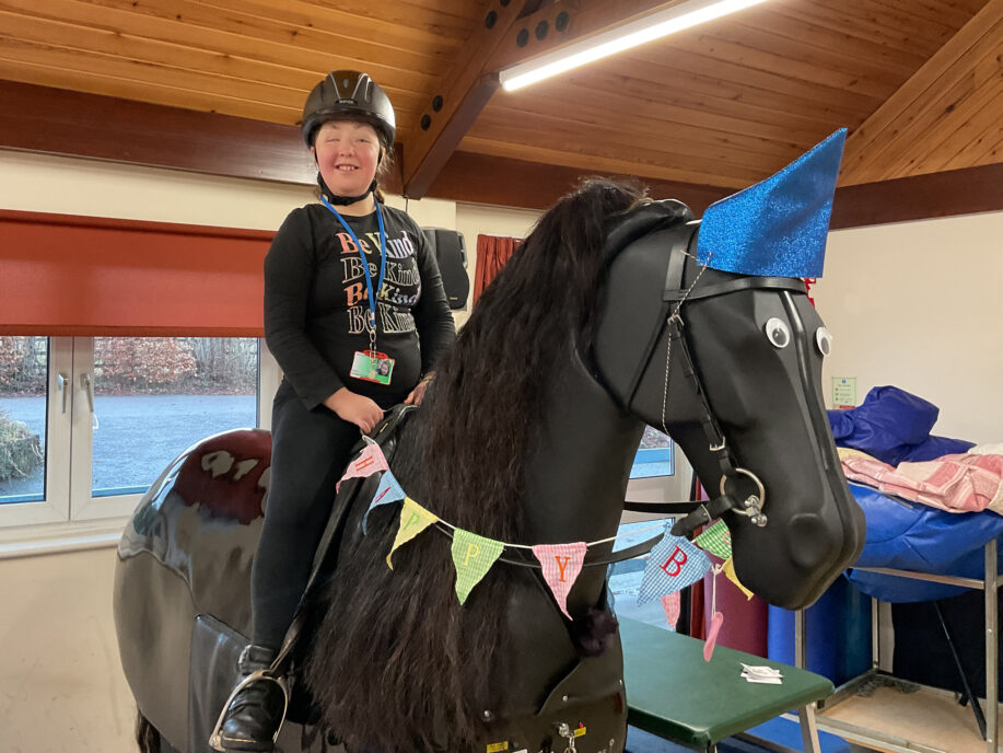 A young woman sat on a mechanical horse. The horse is wearing a party hat and has birthday bunting drapped on it.