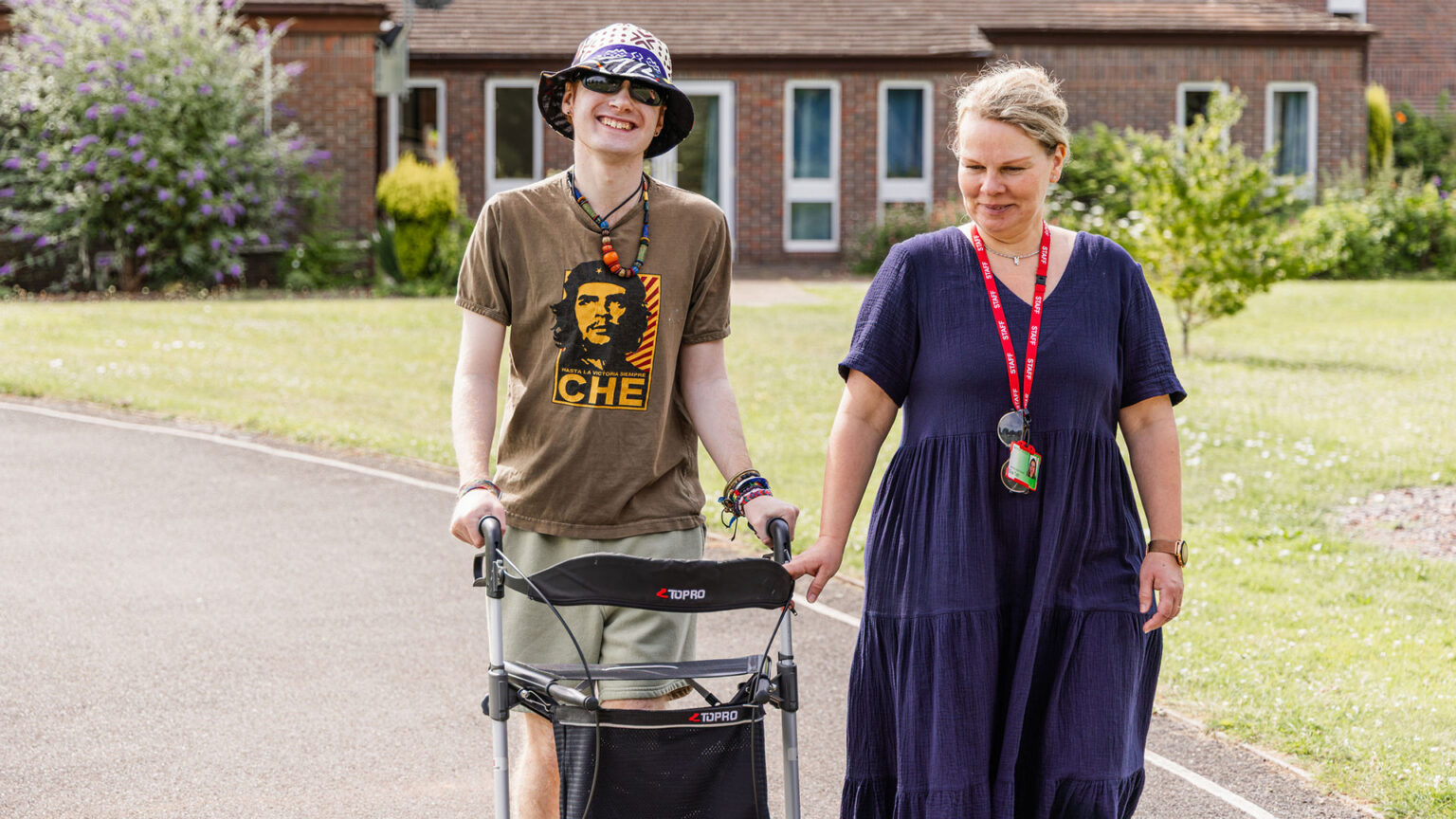 A young man walking with a walker. A lady is walking with him. They're smiling