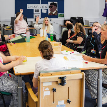 a classroom scene with students and teachers sat around with their arms raised