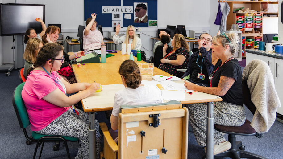 a classroom scene with students and teachers sat around with their arms raised