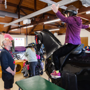 A person is sat on a mechanical horse with their arms raised to the side. A man watches them.