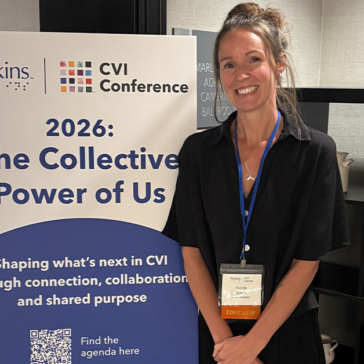 A woman stood next to a banner that says CVI Conference, The Collective Power of Us. She's facing the camera and smiling.
